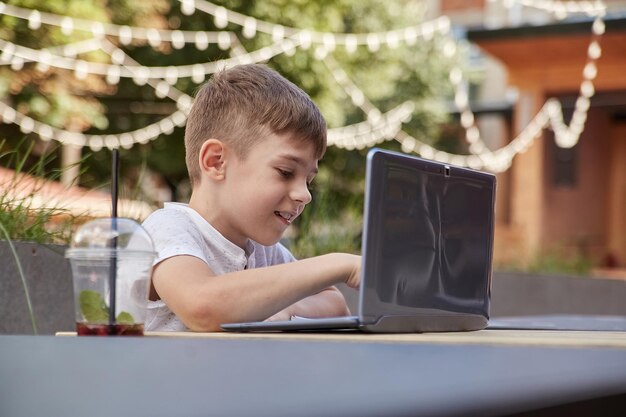 Pequeno estudante caucasiano sorridente senta-se ao ar livre no teclado do laptop