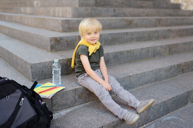 Pequeño estudiante sentado en la escalera cerca del edificio de la escuela.