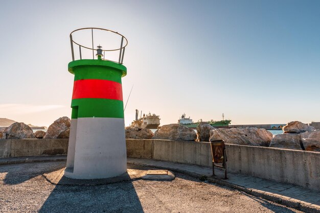 Pequeño faro costero con el sol detrás con grandes barcos al fondo Motril Andalucía España