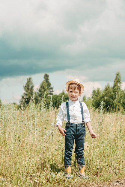 Pequeno fazendeiro em um campo de verão, menino bonitinho com um chapéu de palha. menino com uma flor fica em um campo. rapaz com botas de borracha e uma camisa branca. estilo sertanejo