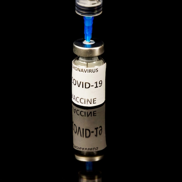 Pequeño frasco de vacuna con tapa plateada y etiqueta Covid - 19 Vacuna contra el coronavirus, jeringa médica aislada sobre fondo negro brillante. Concepto de vacunación. De cerca. Enfoque selectivo. Copia espacio