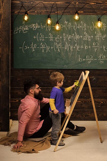 Pequeño genio aprendiendo matemáticas. Maestro o padre ayudando al niño a resolver la ecuación en la pizarra. Chico barbudo en camisa rosa sentada en el suelo