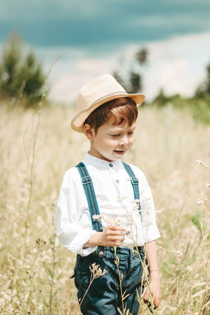 Pequeño granjero en un campo de verano, niño lindo con un sombrero de paja. niño con una flor se encuentra en un campo. chico con botas de goma y una camisa blanca. Estilo rústico