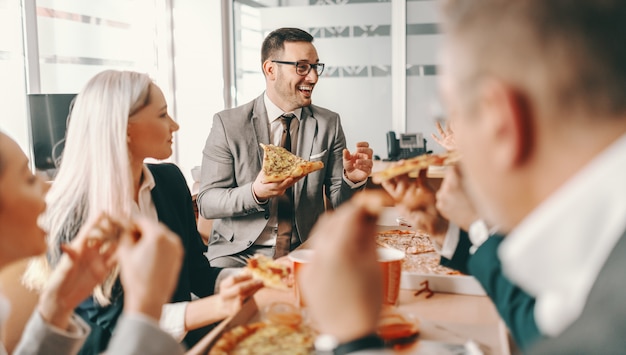 Pequeño grupo de colegas felices en ropa formal charlando y comiendo pizza juntos para el almuerzo. Las grandes cosas en los negocios nunca las hace una sola persona, las hace un equipo de personas.