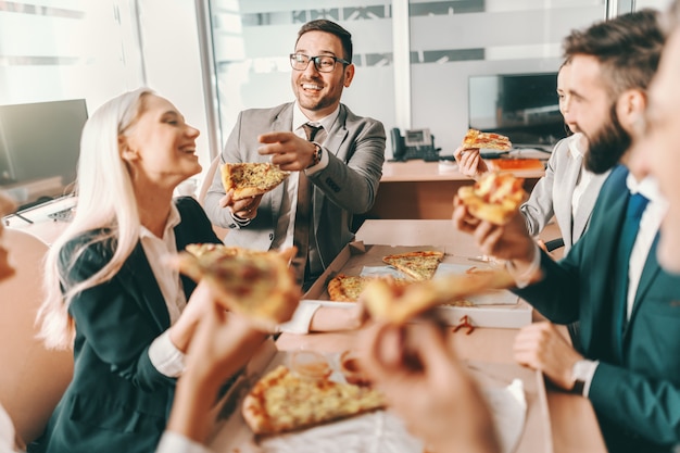 Pequeño grupo de colegas felices en ropa formal charlando y comiendo pizza juntos para el almuerzo. El talento gana partidos, el trabajo en equipo tut gana campeonatos.