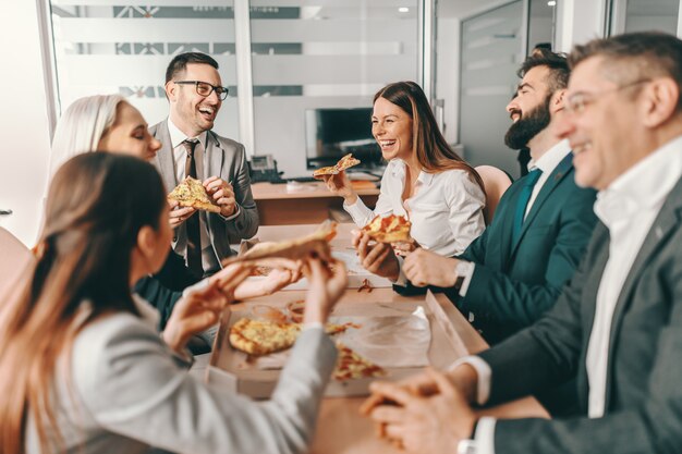 Pequeno grupo de colegas felizes com roupa formal, conversando e comendo pizza juntos no almoço.