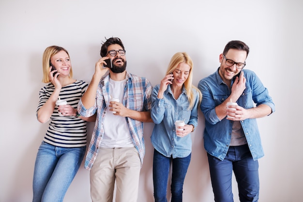 Pequeno grupo de jovens empresários segurando café para ir e telefones inteligentes enquanto conversava e ria. no fundo enquanto parede. arranque o conceito do negócio.