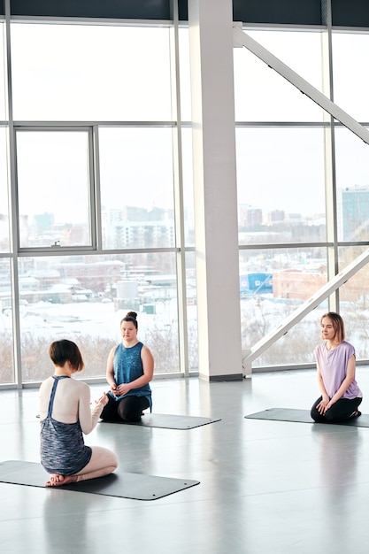 Pequeno grupo de mulheres jovens sentadas em colchonetes em frente ao instrutor de ioga e condicionamento físico explicando-lhes as regras de treinamento na academia