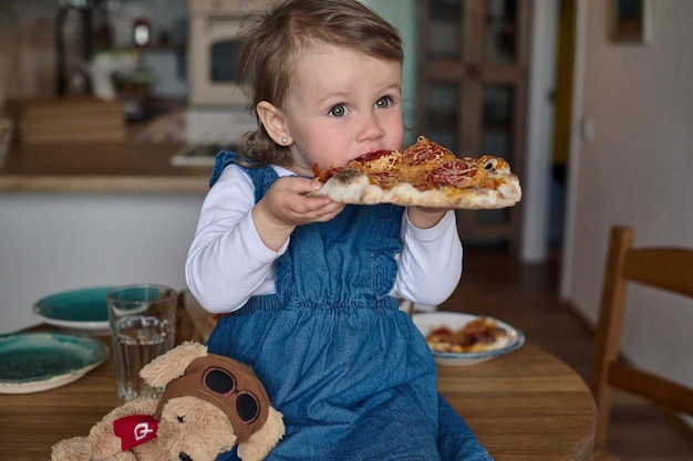 Pequeño y hermoso bebé comiendo pizza sentado en la mesa en la cocina Concepto de familia feliz