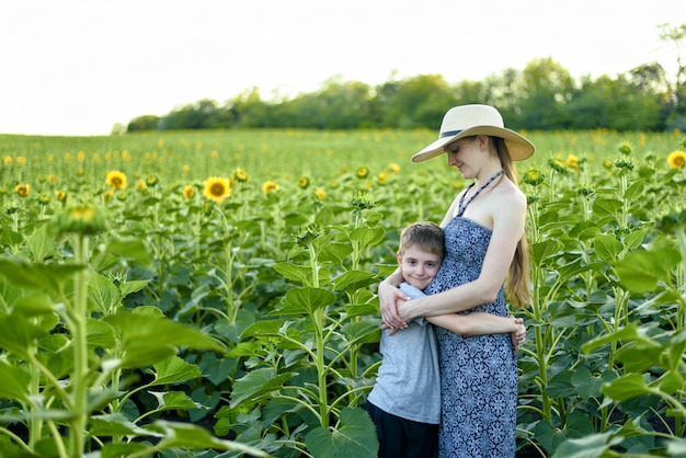 Pequeño hijo abraza a la madre embarazada de pie en un campo de girasoles florecientes