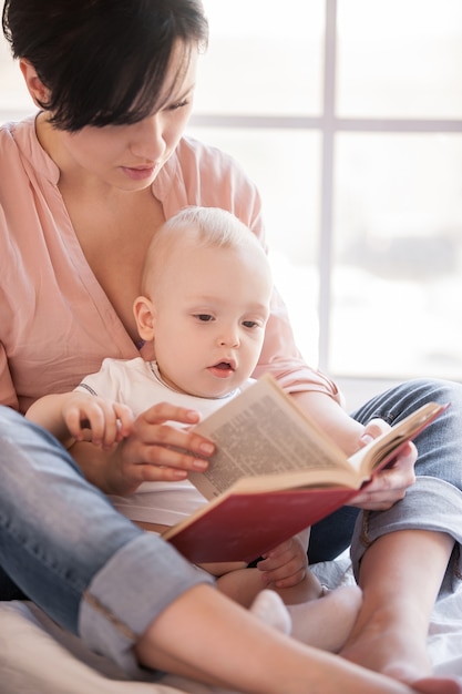 Pequeno leitor ávido. Jovem mãe segurando um bebê e lendo um livro