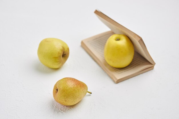 Pequeño libro abierto, manzanas y peras se encuentran sobre una superficie blanca