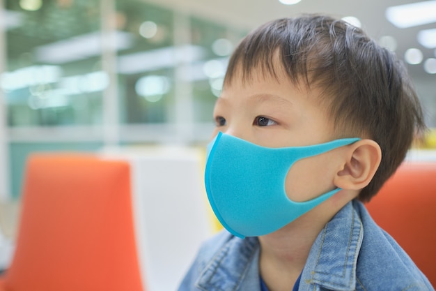 Pequeño y lindo asiático de 3 a 4 años de edad, niño pequeño niño con máscara médica protectora contra la contaminación atmosférica PM 2.5, niño sentado en el sofá esperando ver al médico