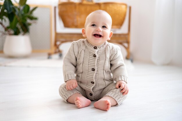 Pequeño y lindo bebé con un traje de punto de lana aprendiendo a sentarse en el suelo en una luminosa sala de estar bebé sonriendo jugando al desarrollo temprano de los niños