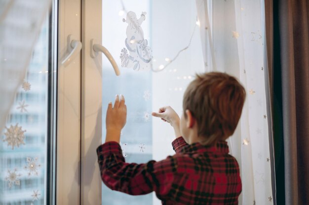 Pequeño y lindo niño caucásico decorando la ventana con pegatinas navideñas