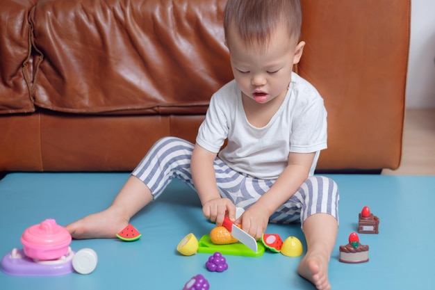 Foto pequeno menino asiático sentado no chão em casa se divertindo brincando sozinho com brinquedos de cozinha