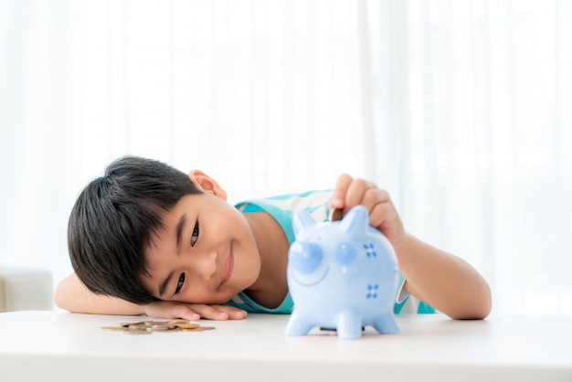 El pequeño muchacho asiático inserta una moneda en la hucha azul en la tabla blanca en la sala de estar en casa para el ahorro del niño.