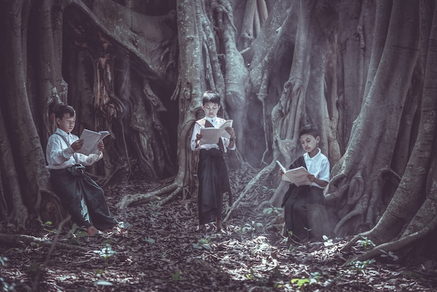 Pequeño muchacho asiático que lee un libro debajo del árbol grande en áreas rurales