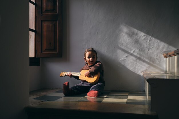 Pequeno músico em roupas casuais sentado no chão de azulejos enquanto toca um instrumento musical em casa