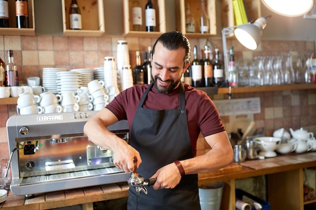 Pequeno negócio, pessoas e conceito de serviço - homem feliz ou garçom de avental com suporte e manipulador preparando expresso em uma cafeteria