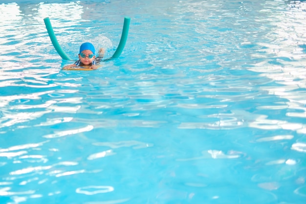 Pequeño niño asiático lindo del niño pequeño que patea los pies en traje de baño usar gafas de natación usar fideos de piscina y kickboard aprender a nadar en la piscina cubierta