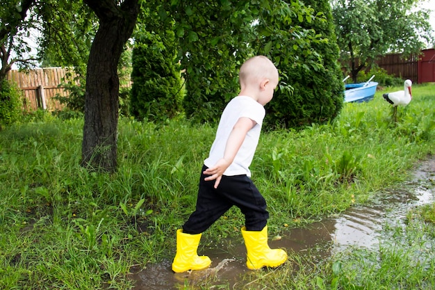 Un pequeño niño calvo con botas amarillas corre por el campo a través de charcos al aire libre