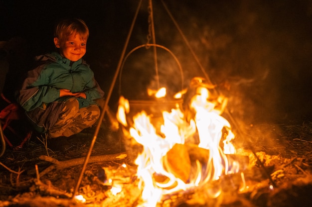 El pequeño niño de cuatro años en una caminata en el campamento está viendo cómo la tetera está en la fogata en la noche. hoguera con una llama ardiente en la oscuridad y gente feliz