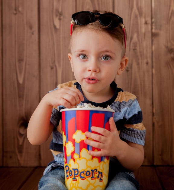 Pequeño niño lindo bebé 2-3 años, gafas de cine 3d con cubo para palomitas de maíz, comiendo comida rápida sobre fondo de madera. Concepto de estilo de vida infantil. Copia espacio