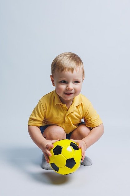 Foto pequeño niño lindo bebé niño 34 años fanático del fútbol en camiseta roja sosteniendo en la mano un balón de fútbol aislado sobre fondo amarillo concepto de estilo de vida de ocio familiar deportivo para niños anuncio de espacio de copia