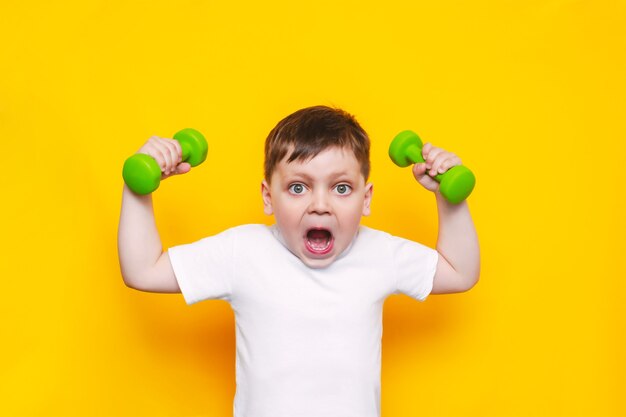 Un pequeño niño lindo con una camiseta blanca con la boca abierta demuestra su fuerza levantando las manos con pesas aisladas en la pared de color amarillo brillante