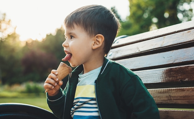 Pequeño niño morena está comiendo helado en el banco del parque mientras mira en algún lugar en un día soleado de verano