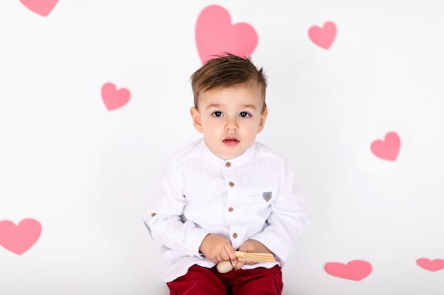 Pequeño niño pequeño en camisa blanca y trouses rojos en el piso blanco con corazones rosados en el día de San Valentín