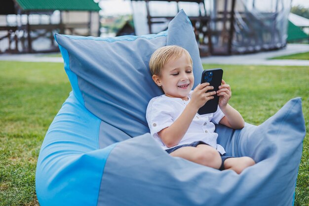 Un pequeño niño rubio de 5 años se sienta en una silla en la calle y mira el teléfono Un niño moderno con un aparato