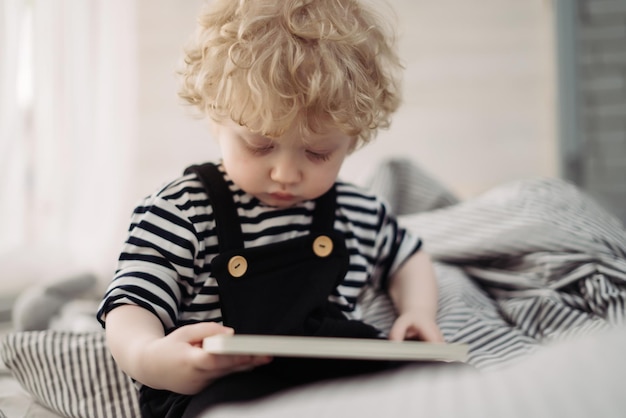 Un pequeño niño rubio de pelo rizado se sienta en la cama y lee un libro.