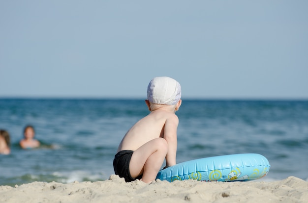 Pequeño niño rubio sentado en la orilla del mar con un círculo de natación con la espalda