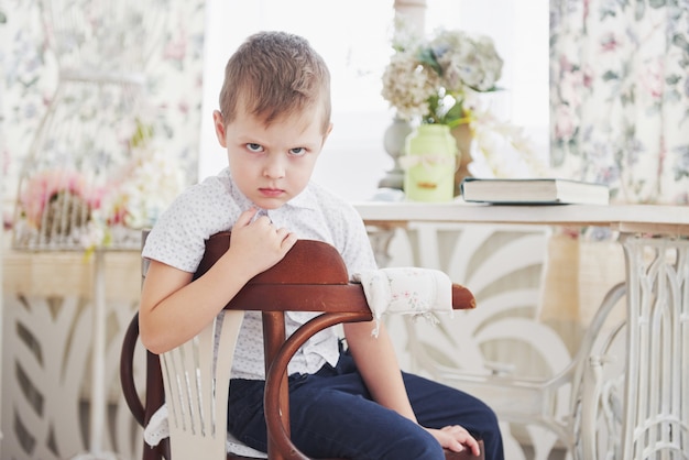 Pequeño niño triste sentado en la silla de madera en la habitación