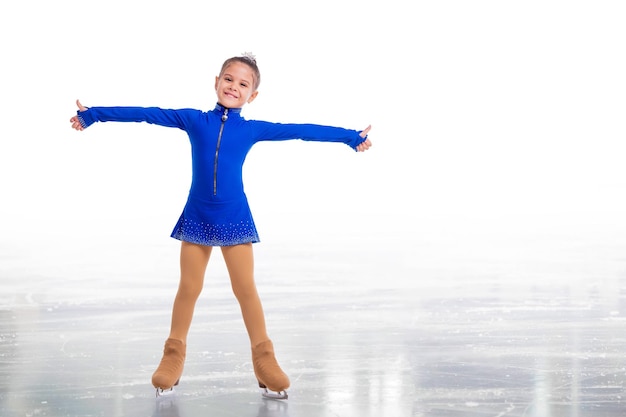 Un pequeño patinador joven posando con un vestido de entrenamiento azul sobre hielo mostrando un gesto de éxito aislado de fondo blanco