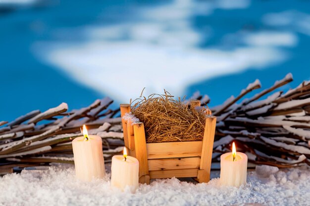 Pequeño pesebre de madera lleno de paja en la nieve y velas alrededor y un cielo azul