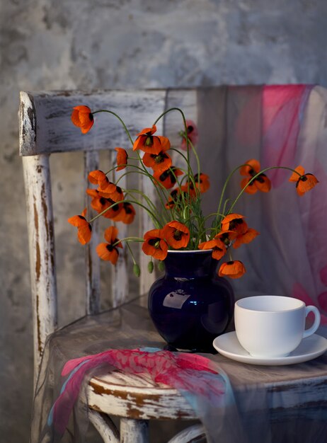 Un pequeño ramo de amapolas rojas en florero azul en silla vintage. Amapolas y taza de café.