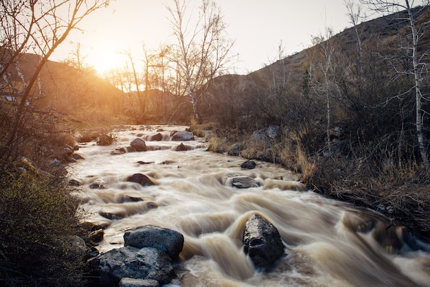 Pequeño río de montaña rodeado de bancos rocosos contra los cerros y el atardecer. Hierba seca y árboles sin hojas en la orilla. Principios de la primavera, la nieve se derrite. Fondo natural.