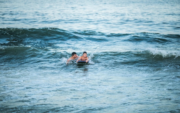 Pequeno surfista aprende a andar de prancha de surf na onda do mar Pai com filho brinca no verão oceano aprendendo a surfar Menino nadar na prancha de surf