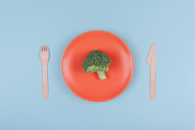 Pequeño trozo de brócoli fresco en plato naranja con tenedor y cuchillo de madera.