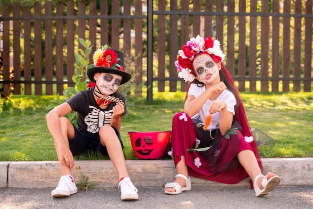 Pequeños amigos divertidos en disfraces de halloween sentados por la carretera frente a la cámara contra la puerta de madera y divirtiéndose y dulces