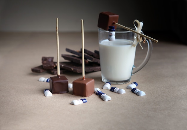Pequenos bonecos de neve perto de cubos de chocolate em um palito com copo de leite e cacau aromático e chocolate em fundo marrom, close-up