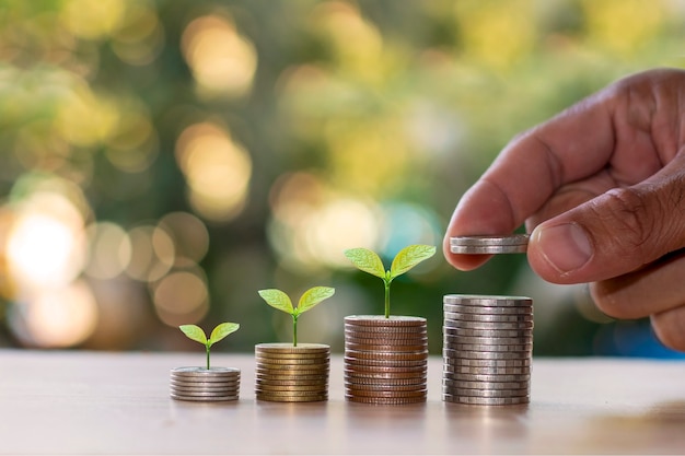 Foto pequeños montones de monedas con pequeñas plantas sobre ellos y una mano recogiendo una moneda.