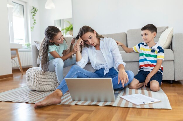 Pequeños niños traviesos que hacen ruido y distraen a la madre freelancer tratando de concentrarse en la computadora portátil y apretando la cabeza trabajando en la mesa en la sala de estar iluminada