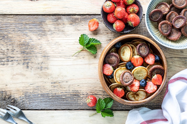 Pequeños panqueques de cereales y mini panqueques de chocolate en un recipiente de madera con miel y fresas sobre un fondo de madera. Vista superior. Copia espacio Comida de moda