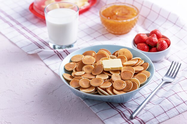 Pequeños panqueques de cereales con una rodaja de mantequilla, fresas, miel y vaso de leche en blanco