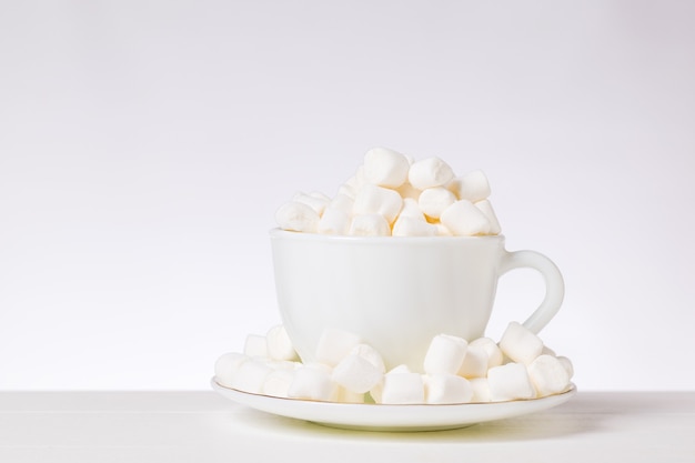 Pequenos pedaços de marshmallow em uma xícara branca e pires sobre uma mesa branca. Sobremesa deliciosa.