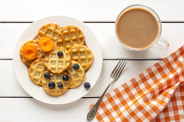 Pequenos waffles redondos com frutas e café sobre fundo branco de madeira, configuração plana